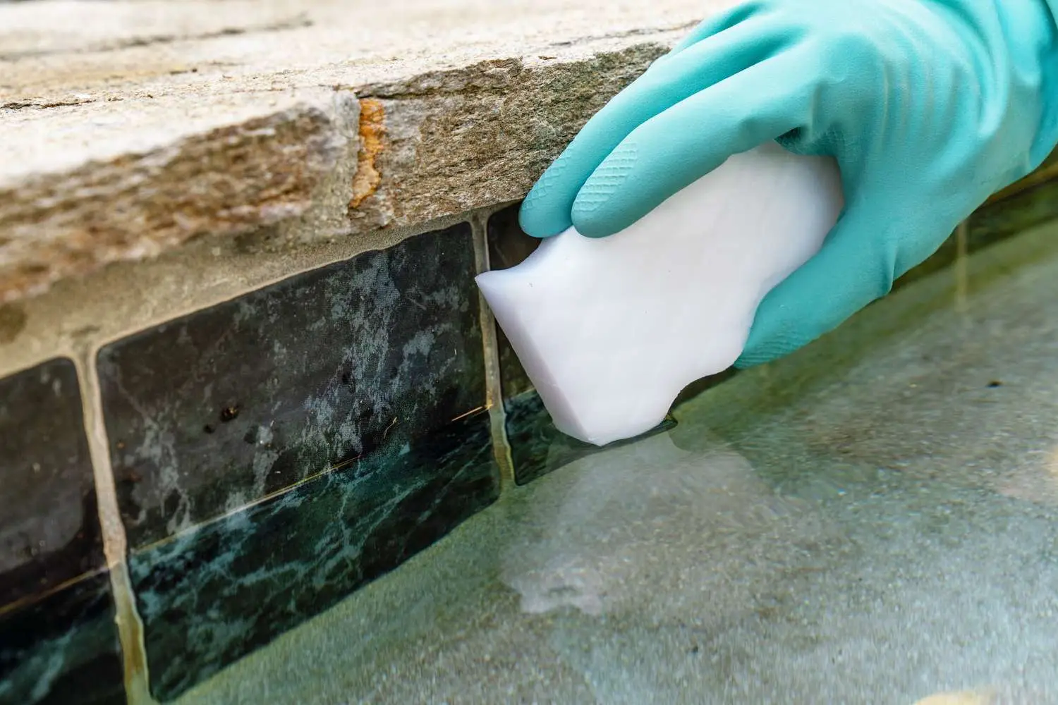 Pool maintenance checklist tasks being done on a North Texas backyard pool Gloved hand scrubs dirty tile grout with a white sponge along a stone edge beside a pool.